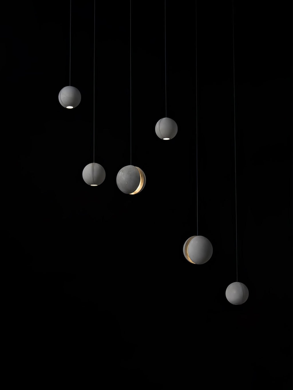 Six Clérachic concrete spheres hang at staggered heights on black, a few with a lit slot and others a light at the base.