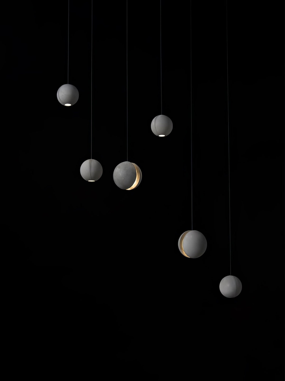 Six Clérachic concrete spheres hang at staggered heights on black, a few with a lit slot and others a light at the base.