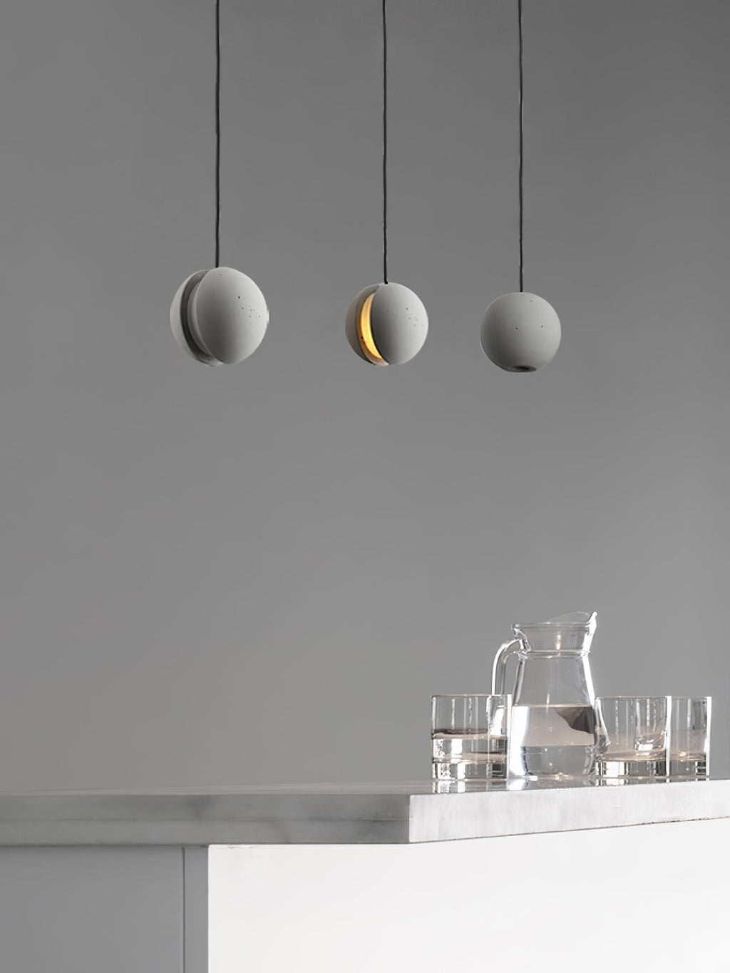 Three Clérachic concrete pendants hang against a mid-grey wall over a counter holding a glass jug and tumblers.