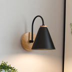 A black Clérachic wall light with a wooden backplate, shown close up with a soft glow on a pale wall and a plant at lower left.