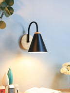 Lit black cone shade with a curved stem and wooden backplate, this Clérachic wall light above a shelf with small wooden ornaments.