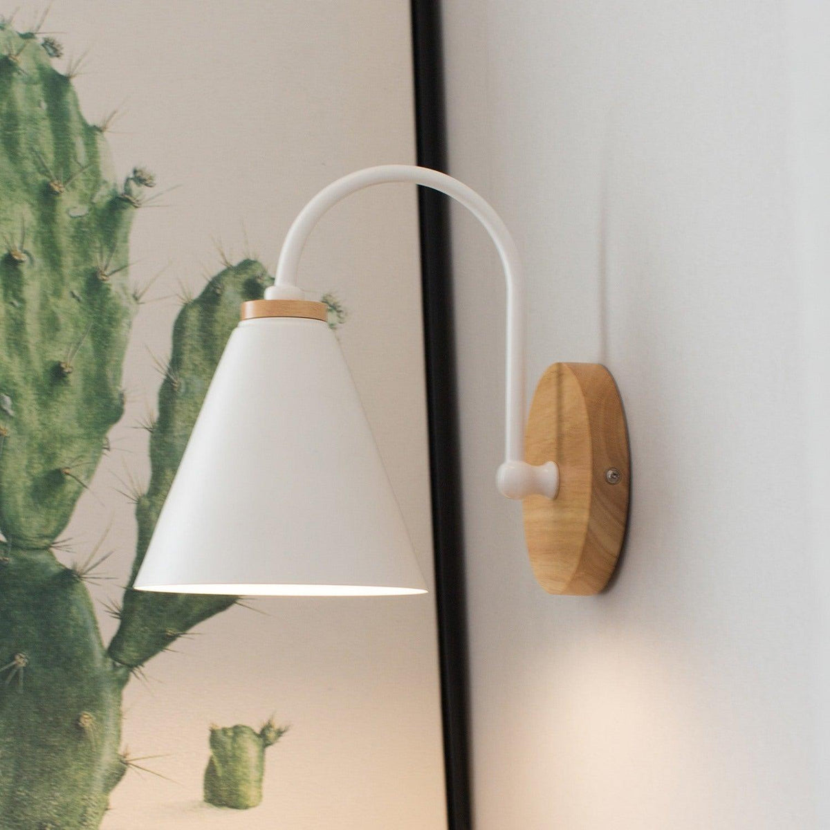 White cone shade on a curved stem, this Clérachic wall light next to a cactus-print picture frame on a grey wall.