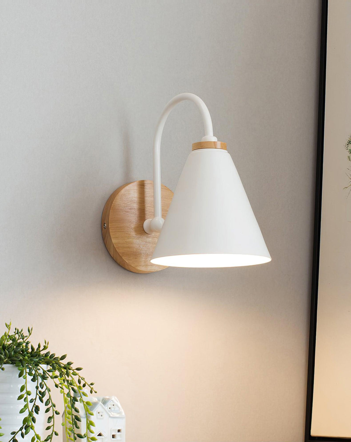A white Clérachic wall light glowing on a grey wall, beside a black picture frame and a trailing plant below.