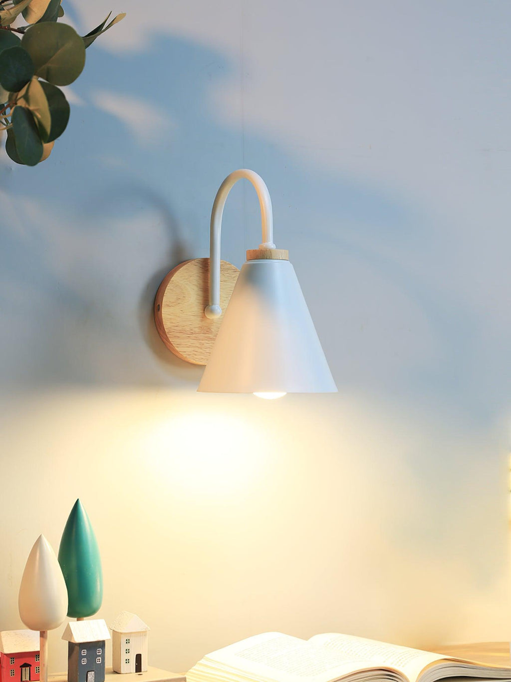 A white Clérachic wall light lit on a wall against a soft blue background, with small wooden houses on a shelf below.