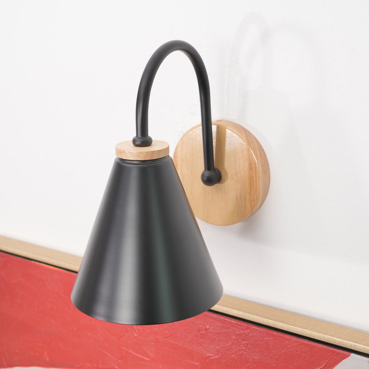 A black Clérachic wall light on a white wall above a bright red picture frame, with its wooden backplate at the right.