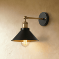 Clérachic wall lamp with a wide black cone shade, brass-toned arm and fittings and a round black backplate, lit by a filament bulb.