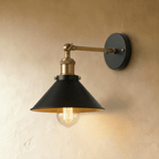 Clérachic wall lamp with a wide black cone shade, brass-toned arm and fittings and a round black backplate, lit by a filament bulb.