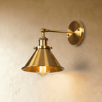 Clérachic wall lamp in brushed brass-tone gold with a wide cone shade and jointed arm, a filament bulb glowing beneath.