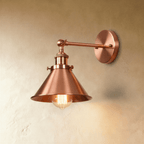 Clérachic wall lamp in a rosy copper finish with a wide cone shade, jointed arm and round backplate, lit by a filament bulb.