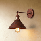 Clérachic wall lamp in a mottled rust-brown aged finish with a wide cone shade and jointed arm, lit by a filament bulb.
