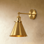 Clérachic wall lamp in bright brushed gold with a narrow cone shade, jointed arm and round backplate on a plaster wall.