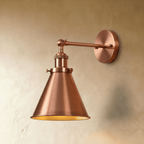 Clérachic wall lamp in a rose copper finish with a narrow cone shade and a glowing gold-toned interior on a plaster wall.