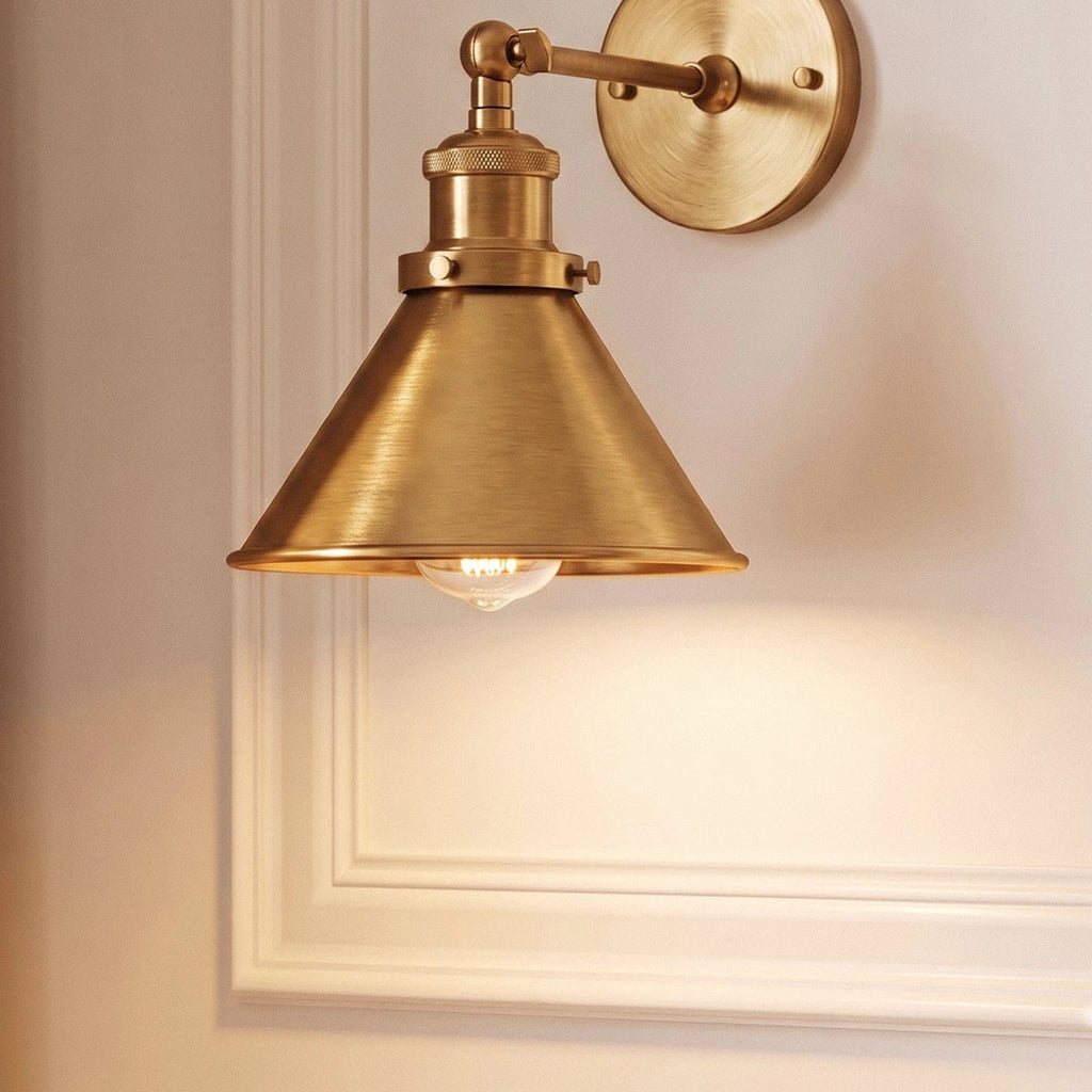 Close view of a Clérachic brushed brass wall lamp: wide cone shade, knurled collar and jointed arm against white moulding.