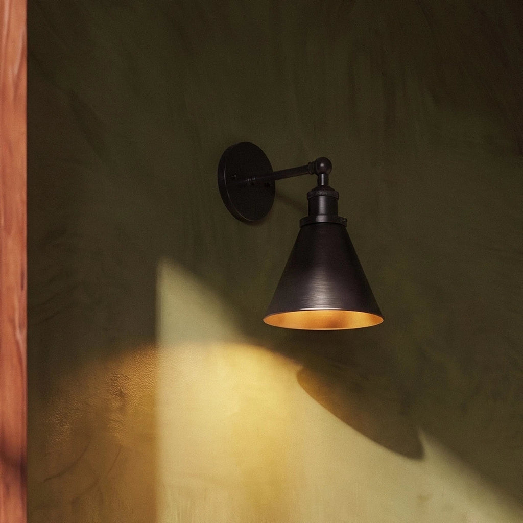 Clérachic dark-finish wall lamp with a narrow cone shade and golden interior casting a pool of light on an olive plaster wall.