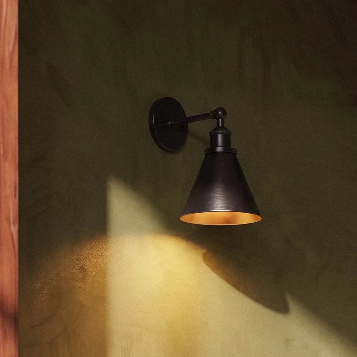 Clérachic dark-finish wall lamp with a narrow cone shade and golden interior casting a pool of light on an olive plaster wall.