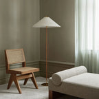 A Clérachic floor lamp with a white conical shade in a sage-grey room, beside a caned wooden chair and a bouclé daybed.