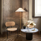 Clérachic floor lamp with a white cone shade in a corner of aged panelled walls, next to a tan armchair and a stone-top round table.