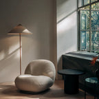 Clérachic floor lamp showing a beige cone shade by tall steel windows, above a cream bouclé chair and a black round side table.