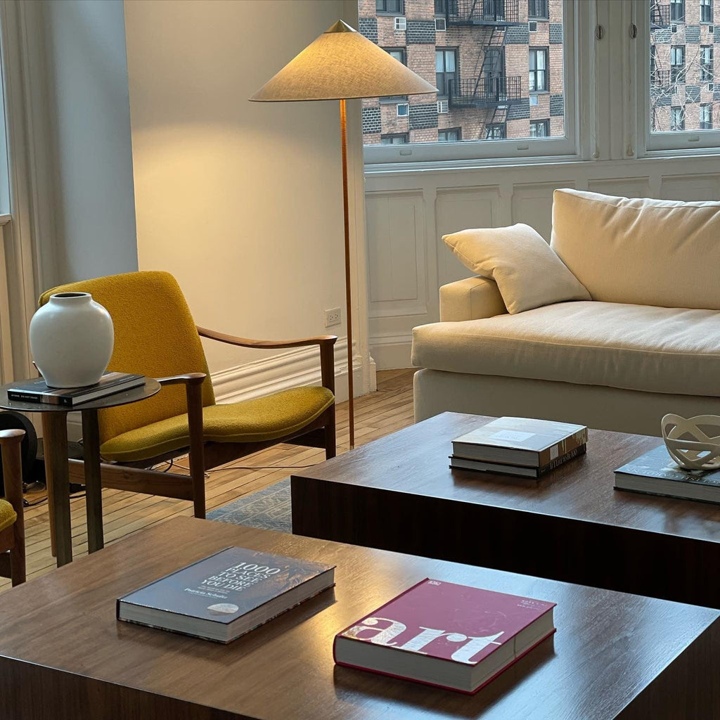 Tall Clérachic floor lamp with a linen-toned cone shade leaning over a mustard armchair and a dark wood coffee table with books.