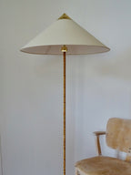 Close view of the Clérachic floor lamp's white cone shade, brass cap and rattan-wrapped stem, with a pale wooden chair beside it.