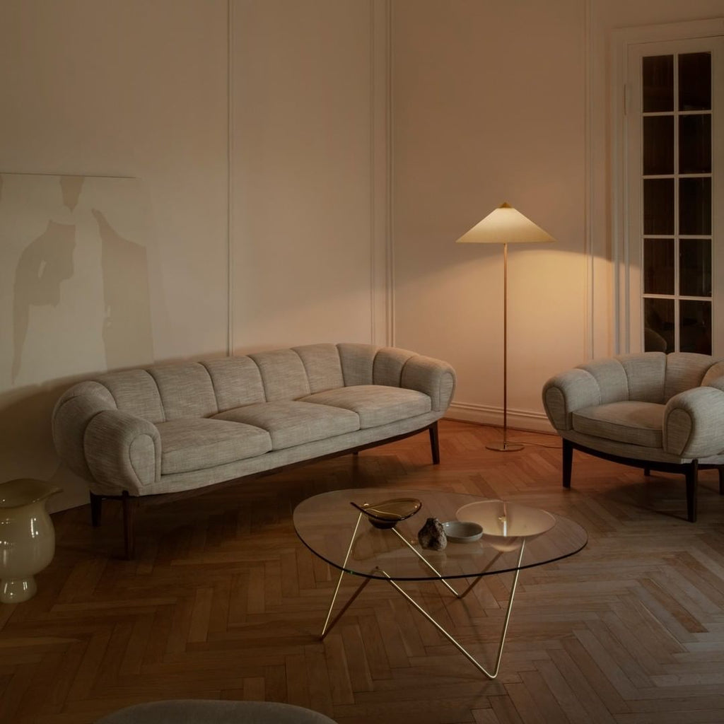 Slim-stemmed Clérachic floor lamp with a pale cone shade lit in a herringbone-floor room, by a grey curved sofa and a glass coffee table.