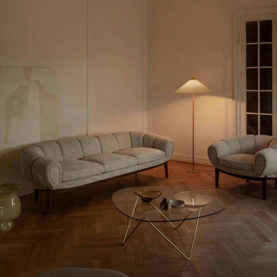 Slim-stemmed Clérachic floor lamp with a pale cone shade lit in a herringbone-floor room, by a grey curved sofa and a glass coffee table.