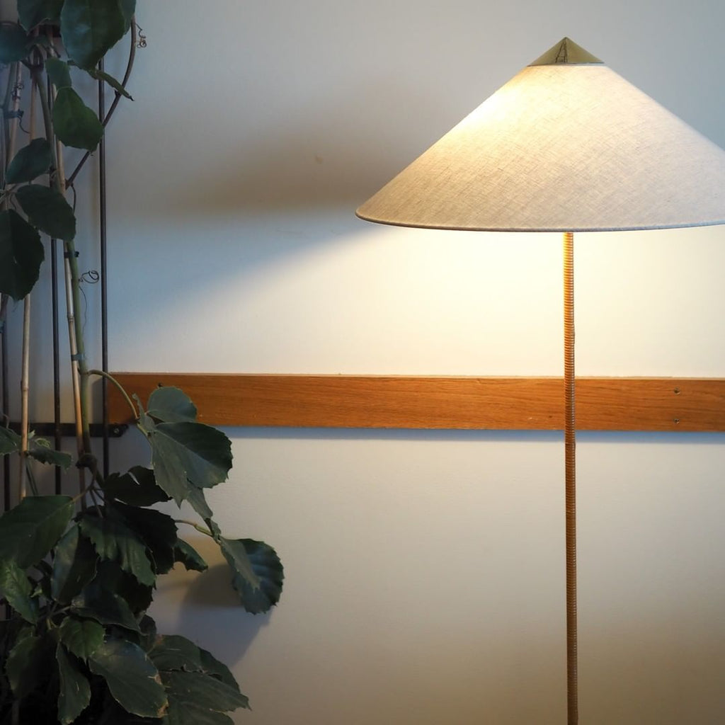Close view of a Clérachic floor lamp with a linen cone shade and brass cap, against a white wall with a wooden rail and a plant.