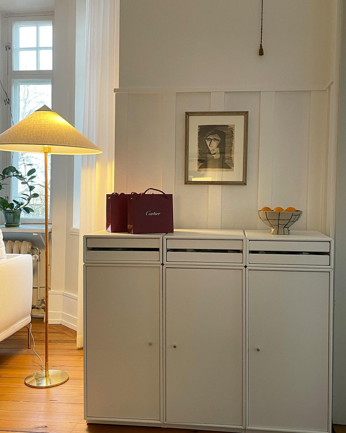 Floor lamp by Clérachic, with a warm linen cone shade over a white sideboard, with a framed portrait on the wall behind.