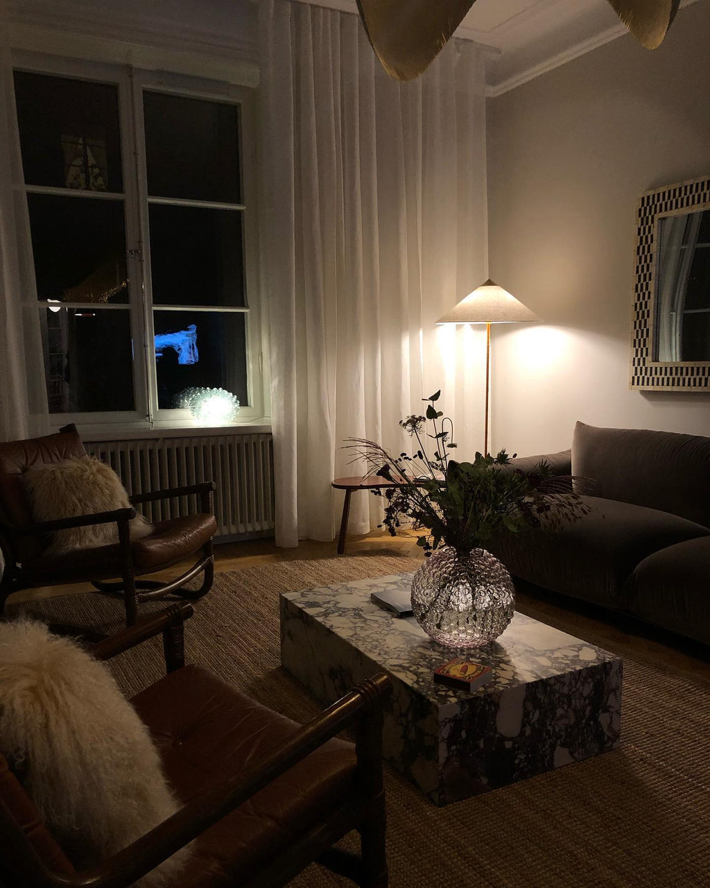 A Clérachic floor lamp glowing softly in a dim evening room, beyond a marble coffee table, a leather chair and a dark sofa.