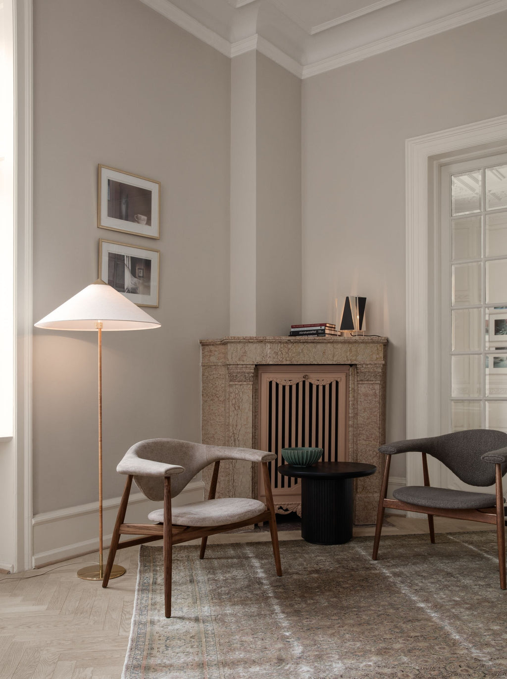 Tall Clérachic floor lamp with a white cone shade in a corner with framed prints, above two grey chairs and a small black table.