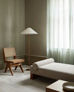 A Clérachic floor lamp with a white cone shade in a sage-green room, beside a caned wooden chair, a daybed and a sheer curtain.