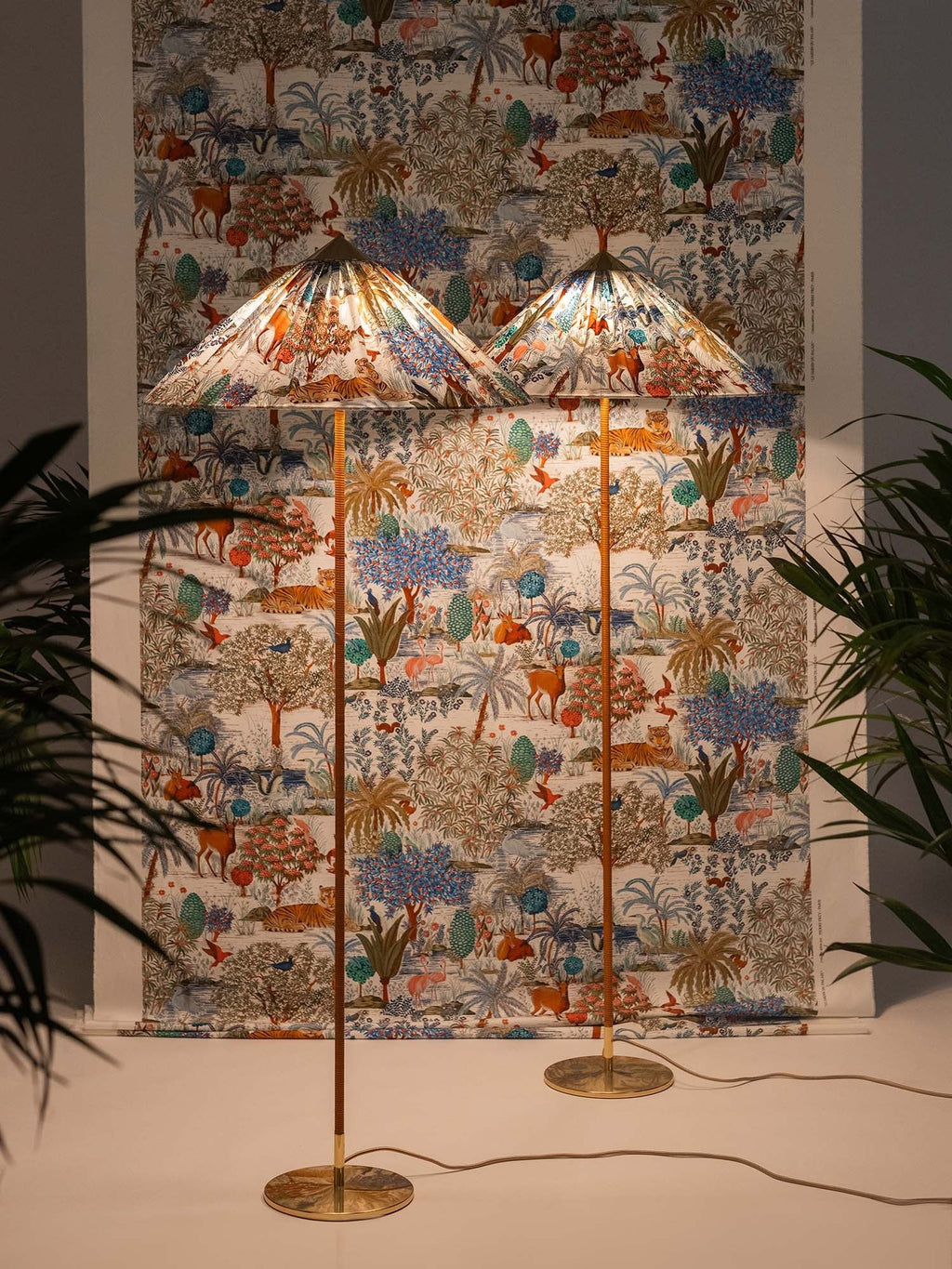Two Clérachic floor lamps with printed fabric cone shades against a matching wall hanging, with palms at the edges.