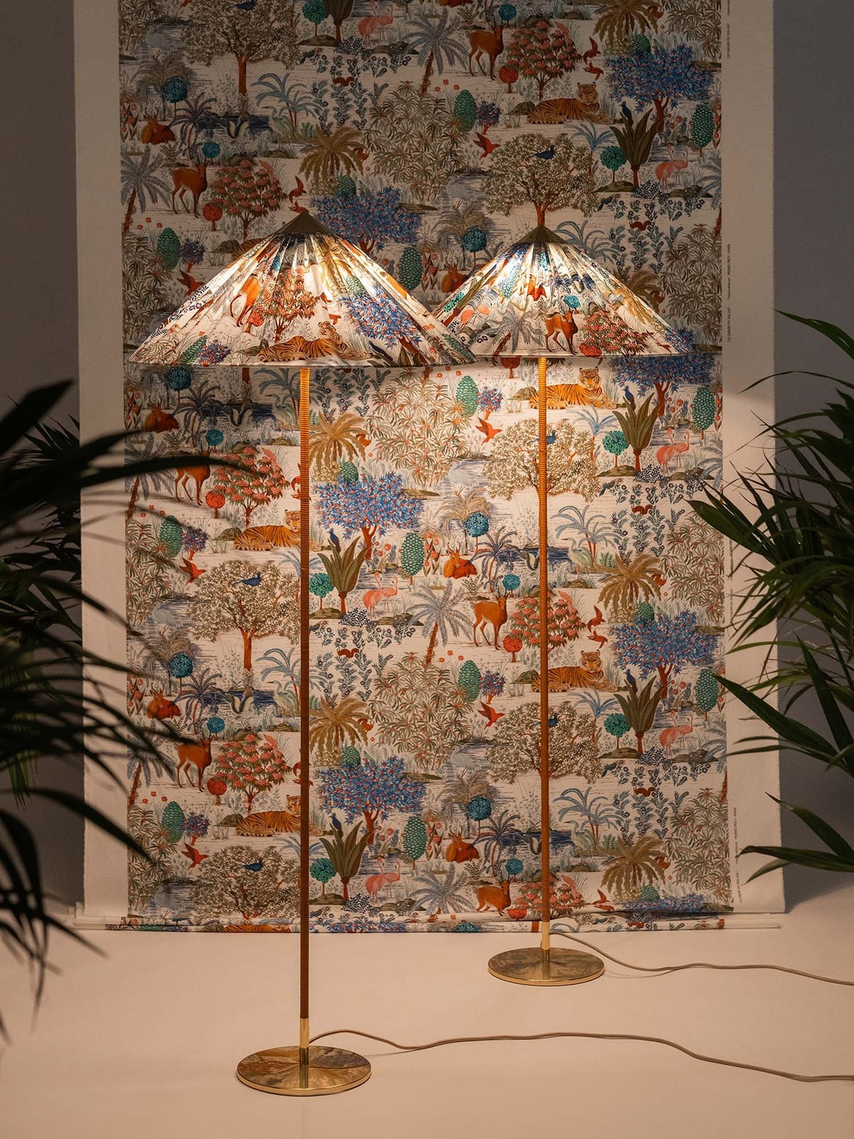 Two Clérachic floor lamps with printed fabric cone shades against a matching wall hanging, with palms at the edges.