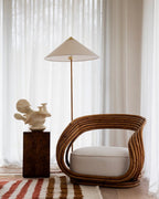 Floor lamp by Clérachic, with a white cone shade behind a curved rattan armchair, with a sculpture on a dark plinth and sheer curtains.