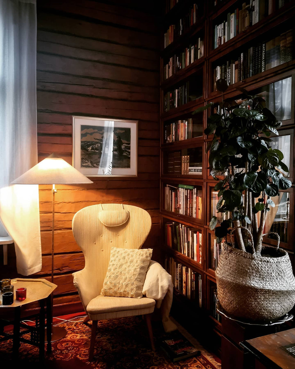 A Clérachic floor lamp lit beside a cream wingback chair in a log-walled reading corner with full bookshelves.