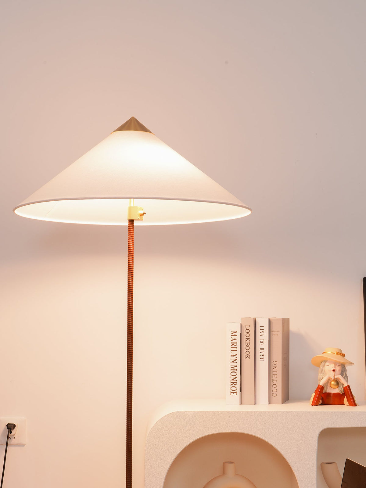 Tall Clérachic floor lamp with a lit white cone shade and brass cap, beside a white shelf with books and a small figure.