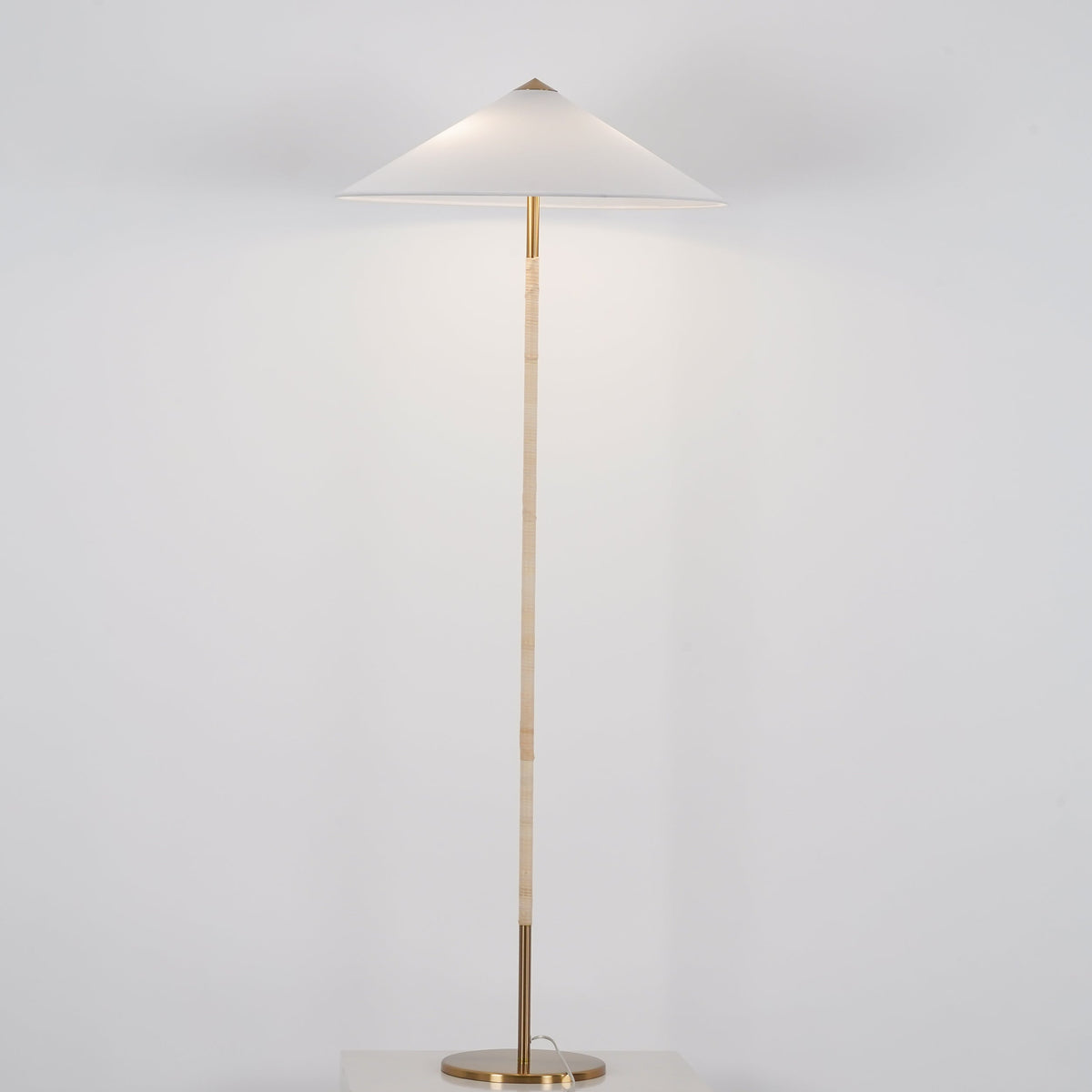Clérachic floor lamp with a white cone shade, a light cane-wrapped stem and a brass base, on a pale grey backdrop.