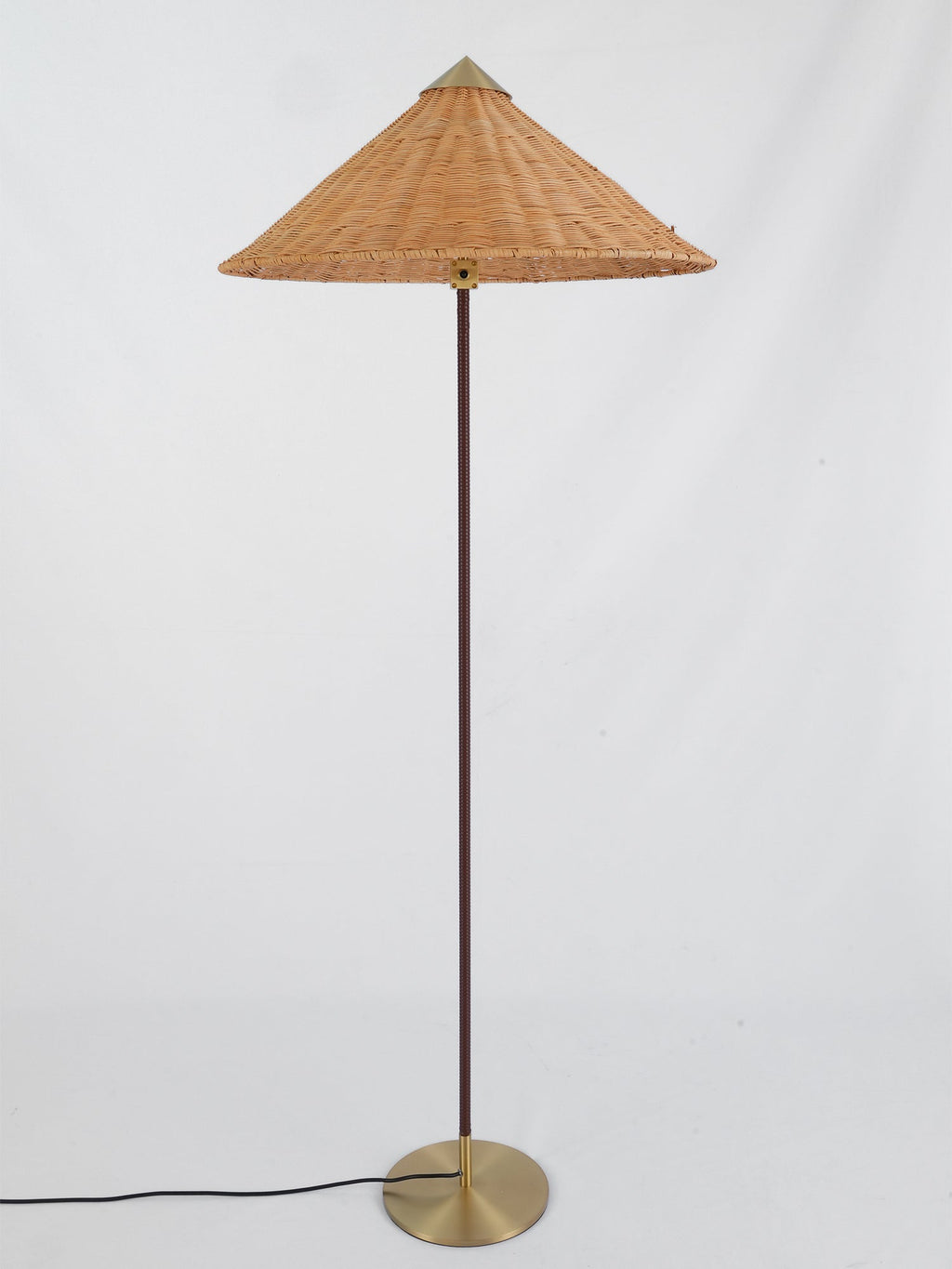 Clérachic floor lamp showing a rattan cone shade, a dark stem and a brass base, with its cord running to the left, on pale grey.