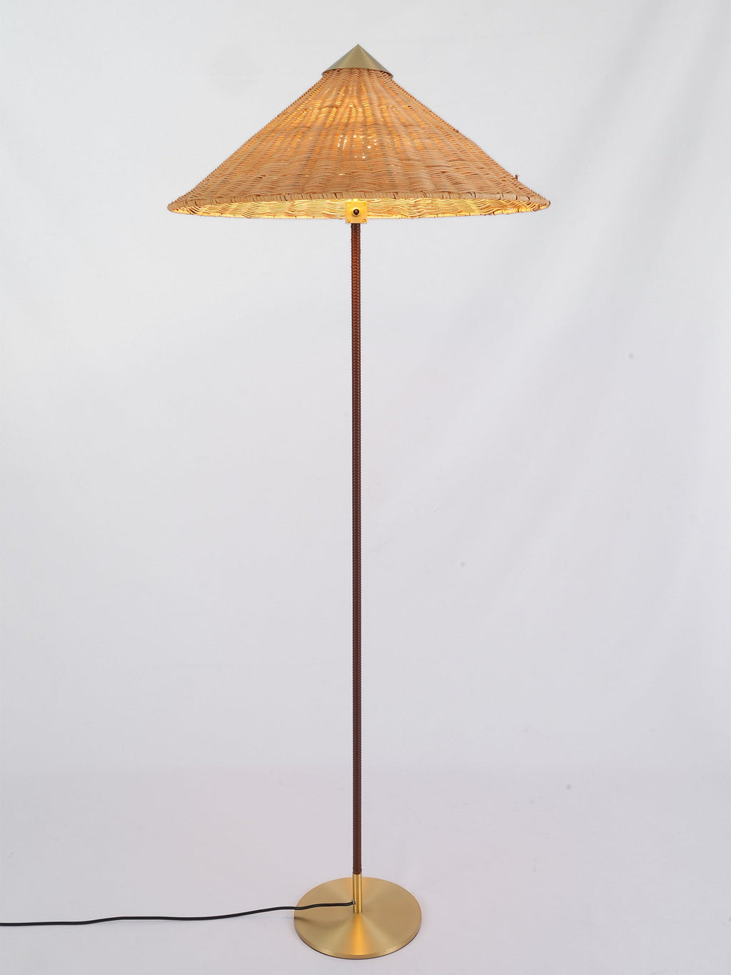 Clérachic floor lamp with a glowing rattan cone shade, a dark stem and a brass base, on a pale grey backdrop.