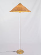 Clérachic floor lamp with a glowing rattan cone shade, a dark stem and a brass base, on a pale grey backdrop.