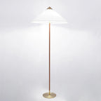 Clérachic floor lamp with a bright white cone shade casting light on a pale backdrop, its stem dark brown over a brass base.