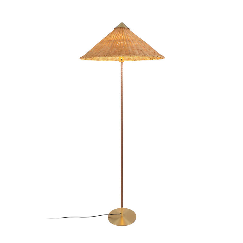 Clérachic floor lamp with a glowing woven rattan cone shade, a dark stem and a black cord trailing left, on white.