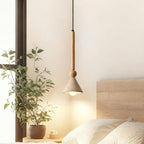 Hung by a window with a leafy plant, a travertine cone pendant on a light wooden stem lights a bed with a pale headboard, from Clérachic.