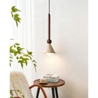 Above a round side table with books and a cup, a travertine cone pendant on a walnut stem hangs in front of a white wall, from Clérachic.