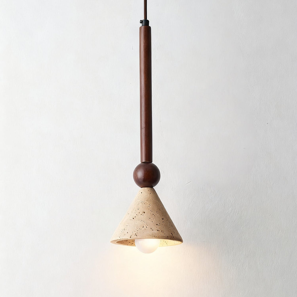 A travertine cone pendant on a walnut stem and a round bead, its warm bulb lit, hangs against a plain pale wall, from Clérachic.