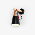 Black conical metal shade hung from a wooden bar by a tan leather strap, this Clérachic wall light on a plain pale backdrop.