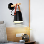 Close view of a black Clérachic wall light over a pale wood headboard, with a glass vase and yellow stems on the bedside table.
