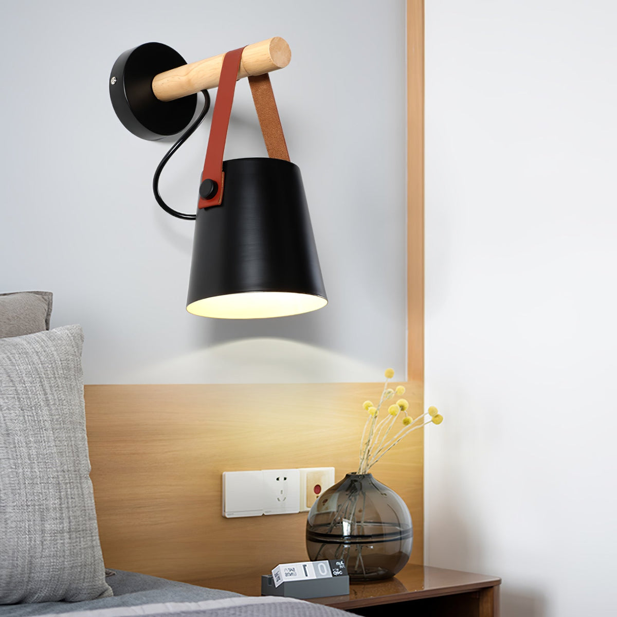 Close view of a black Clérachic wall light over a pale wood headboard, with a glass vase and yellow stems on the bedside table.