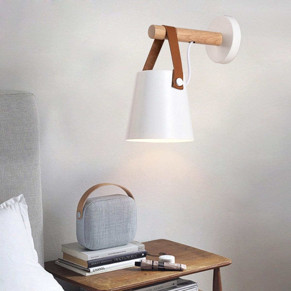 White Clérachic wall light above a wooden bedside table holding a grey fabric speaker and a small stack of books.