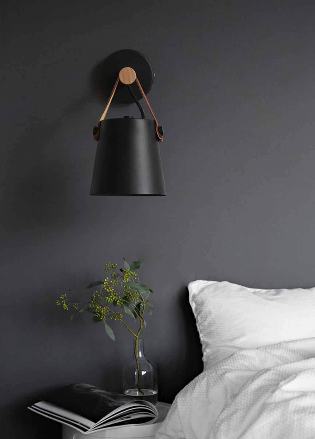 Black Clérachic wall light on a charcoal wall above a bed with white linen, a glass vase of leaves and a magazine.